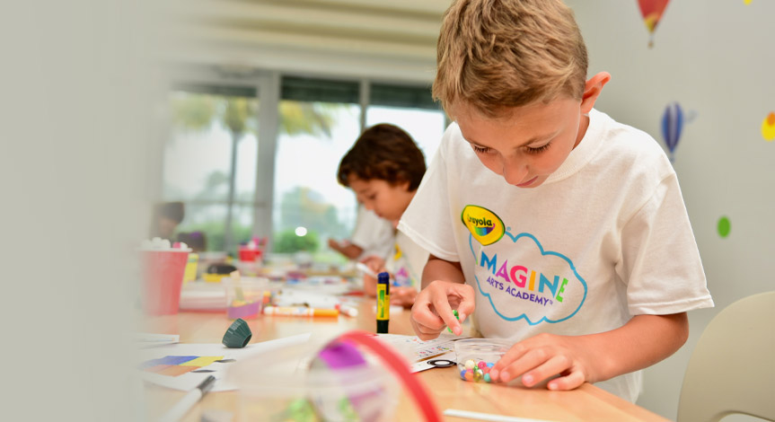 Blonde boy wearing a Crayola imagine arts academy tshirt coloring in an art class