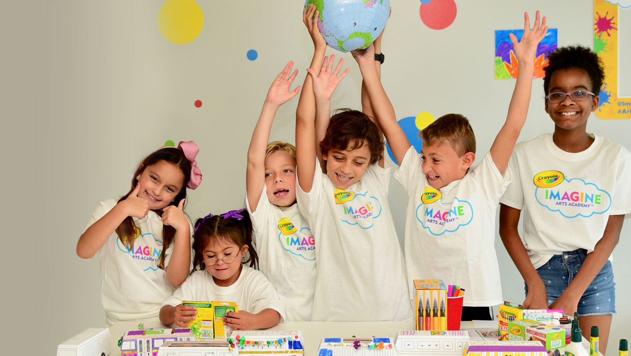 Kids around a desk with their finished art projects. 2 boys are holding a globe they made in class