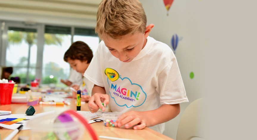 Blonde boy wearing a Crayola imagine arts academy tshirt coloring in an art class